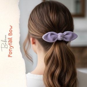 Könnte beinhalten: Eine hellviolette Stoffschleife für den Pferdeschwanz mit einem geknoteten Mittelteil. Die Schleife ist an einem Haargummi befestigt und sitzt in einem tiefen Pferdeschwanz. Der Text Babinaa Ponytail Bow steht vertikal auf der linken Seite.