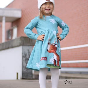 Könnte beinhalten: Ein türkisfarbenes Langarmkleid mit weißem Schneeflockenmuster und einer Grafik eines braunen Rentiers. Das Kleid hat eine geraffte Taille und ein kleines rechteckiges Etikett mit dem Text EINZIGARTIG auf dem Rockteil.