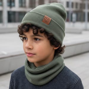 Könnte beinhalten: Eine salbeigrüne, gerippte Strickmütze mit umgeschlagenem Saum und einem kleinen, quadratischen braunen Lederaufnäher mit eingeprägtem Smiley-Gesicht, kombiniert mit einem passenden salbeigrünen, gerippten Schlauchschal über einem dunkelgrauen Pullover.