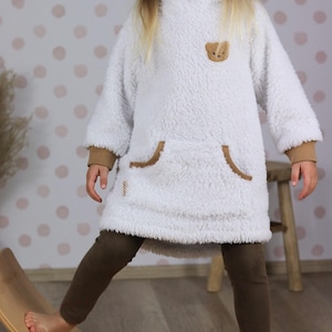 Könnte beinhalten: Ein weißes, langärmeliges Fleece-Pulloverkleid mit einem braunen Bärenaufnäher auf der Brust, einer Kängurutasche mit brauner Umrandung und braunen Rippbündchen. Das Kleidungsstück wird mit einer einfarbigen braunen Leggings kombiniert.