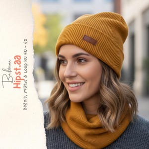 May include: Mustard yellow beanie and matching neck gaiter. The beanie has a brown leatherette patch. The text on the left side of the image reads "Bapinna Hipst.aa Beanie, Mütze & Loop KU 40 - 60". The woman is wearing a gray sweater.