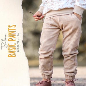 May include: Beige corduroy pants featuring an elastic waistband and ribbed cuffs at the ankles. The text on the left side reads Babinaa BASIC PANTS Größe 92 - 146. The pants have side pockets and a vertical textured pattern throughout the fabric.