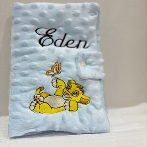 Könnte beinhalten: Eine hellblaue Babydecke mit strukturierter Oberfläche und einem gestickten Bild eines gelben Löwenjungen und eines Schmetterlings. Der Name "Eden" ist in schwarzer Schrift gestickt. Die Decke hat einen Knopfverschluss.