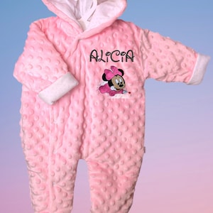 Peut inclure: Combinaison rose pour bébé avec une capuche et des oreilles blanches. La combinaison est brodée d'un motif de Minnie Mouse et du nom "Alicia" sur le devant.