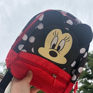 Peut inclure: Un sac à dos noir avec un motif à pois blancs et une grande illustration de Minnie Mouse sur le devant. Le sac comprend une poche avant rouge avec un motif à pois blancs plus petits, une fermeture éclair et deux oreilles noires sur le dessus.