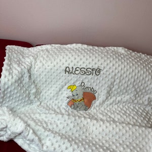 Peut inclure: Une couverture blanche pour bébé avec un éléphant brodé gris avec le nom "Dumbo" et le nom "Alessio" au-dessus.