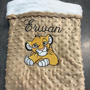 Puede incluir: Una manta de bebé beige con un borde de felpa blanco. La manta presenta una imagen bordada de un cachorro de león de dibujos animados y el nombre "Erwan" en cursiva. La manta tiene un patrón texturizado con puntos.