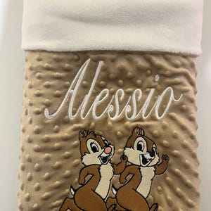 Könnte beinhalten: Eine beige und weiße Babydecke mit dem personalisierten Namen "Alessio", der in Weiß gestickt ist. Die Decke zeigt zwei gestickte Comic-Streifenhörnchen, die rennen.