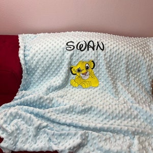 Puede incluir: Una manta de bebé azul claro con un cachorro de león amarillo bordado y el nombre "SWAN" bordado en negro.
