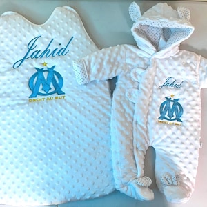 Peut inclure: Turbulette et combinaison à capuche blanches pour bébé avec broderie bleue et dorée. La broderie comprend le nom "Jahid" et un logo avec le texte "Droit au But".