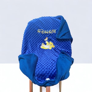 Puede incluir: Una manta para bebé azul y blanca con un personaje de león y el nombre "ADHAM" bordado.