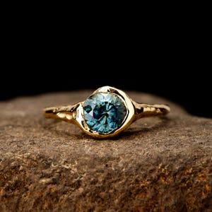 May include: A gold ring featuring a round, faceted blue gemstone set in a textured, organic-shaped metal band. The setting surrounds the stone with a polished gold finish, contrasting with the uneven, hammered appearance of the ring shank.