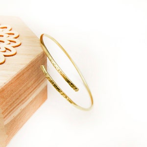 Puede incluir: Un brazalete de oro martillado con un diseño simple y abierto. El brazalete está hecho de una sola pieza de metal que se dobla en un círculo y luego se martilla para crear una superficie texturizada.