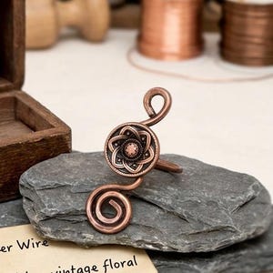 May include: A copper-toned metal ring featuring a central circular element with a raised floral pattern and two spiraling wire ends. The ring is displayed on a dark gray stone, with partial text visible on a paper below reading, er Wire with a vintage floral.