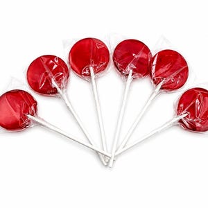 Può includere: Sei lecca-lecca rotondi di colore rosso brillante disposti a ventaglio. Ogni caramella è avvolta singolarmente in una pellicola di plastica trasparente e stropicciata, fissata a un bastoncino di carta bianco, sottile e rigido.