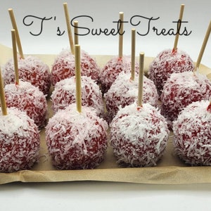 May include: A dozen red candy apples coated in shredded coconut, each with a wooden stick. The apples are arranged on a brown paper-lined tray. The text "T's Sweet Treats" is written in cursive above the apples.