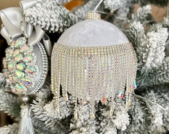 May include: Two spherical Christmas ornaments featuring white velvet surfaces. One ornament is adorned with iridescent teardrop-shaped crystals and a silver ribbon bow. The other features a wide band of dangling crystal fringe trim.