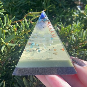May include: A green, clear, and black pyramid-shaped crystal with copper wire and various crystals inside. The pyramid is sitting on a hand.