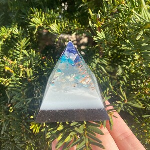 May include: A small white pyramid with blue and green crystals embedded in the resin. The pyramid has a black base and a copper wire wrapped around it.