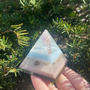 May include: A clear resin pyramid with layers of blue, white, and pink resin. A copper wire coil is embedded in the pyramid. The pyramid is sitting on a hand.