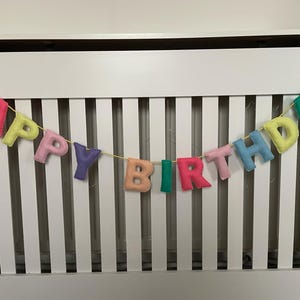 May include: A colorful felt banner with the words "Happy Birthday" strung across a white slatted surface.