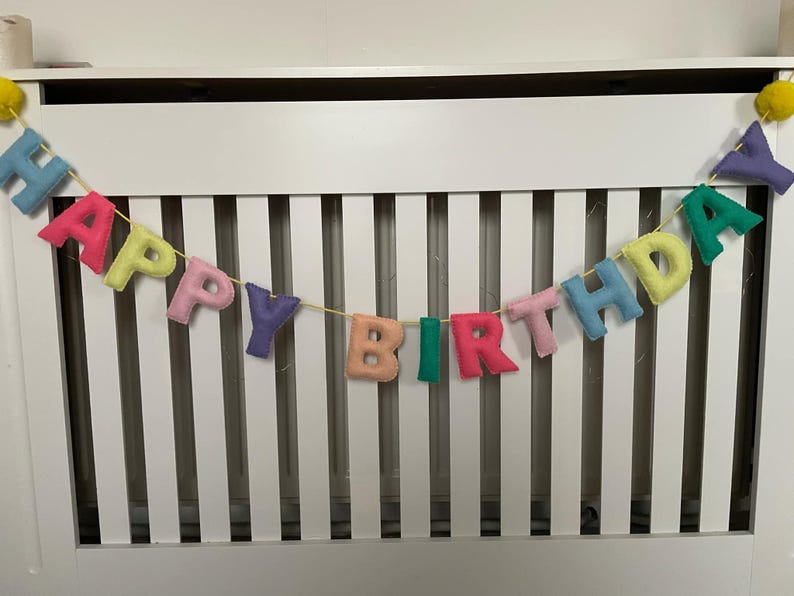 May include: A colorful felt banner with the words "Happy Birthday" strung across a white slatted surface.