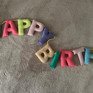 May include: A colorful felt banner that reads "Happy Birthday" with each letter in a different color. The banner is strung together with yellow string.