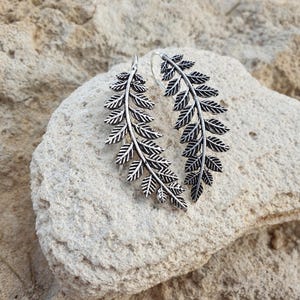 May include: A pair of silver leaf earrings. Each earring has a detailed fern leaf design. The earrings are shown on a light-coloured, textured stone surface.