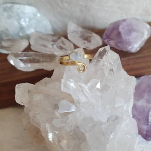 May include: A gold-colored ring with a light blue gemstone and a spiral design. The ring is displayed on a cluster of clear quartz crystals, with other crystals in the background. The ring is a piece of jewelry.