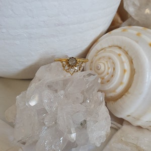 May include: A gold ring with a clear stone set in a delicate floral design. The ring is displayed on a bed of white crystals.