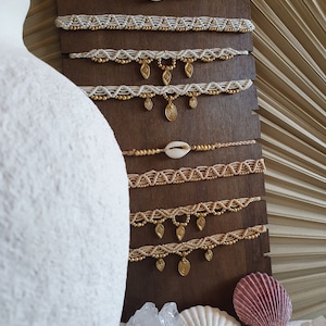 May include: A display of handmade macrame chokers with gold beads and pendants, some featuring seashells. The chokers are displayed on a dark wooden stand, with a white vase and seashells in the foreground.