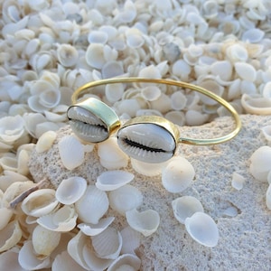 Peut inclure: Un bracelet en or avec deux coquillages blancs attachés. Le bracelet est posé sur un lit de coquillages blancs.