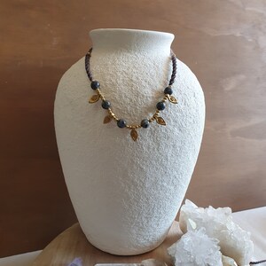 May include: A dark purple and gold necklace with small round beads and leaf-shaped charms. The necklace is displayed on a white ceramic vase and a wooden surface with crystals.