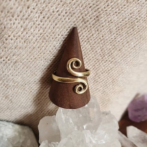 May include: A gold ring featuring a double swirl design. The ring is presented on a brown, cone-shaped stand, set against a backdrop of crystals and a textured, off-white fabric.