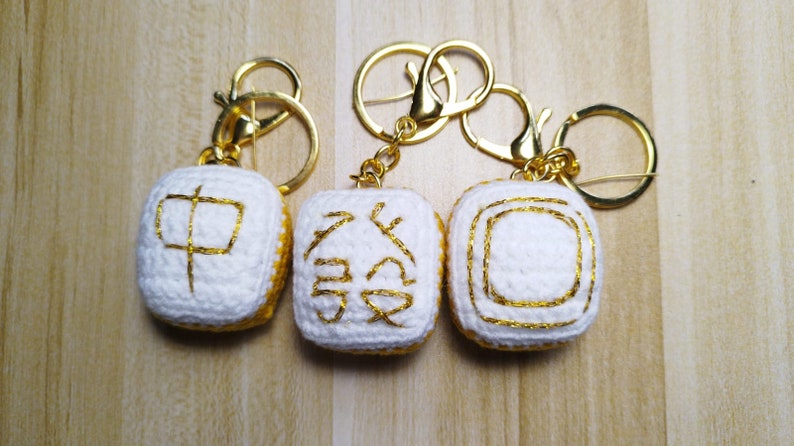 K&ouml;nnte beinhalten: Drei wei&szlig;e geh&auml;kelte Schl&uuml;sselanh&auml;nger mit goldener Stickerei. Die Schl&uuml;sselanh&auml;nger sind quadratisch und zeigen chinesische Schriftzeichen. Die Schl&uuml;sselanh&auml;nger sind an goldenen Schl&uuml;sselringen befestigt.