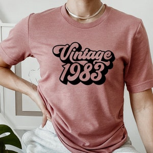 Puede incluir: Una camiseta rosa con un gráfico negro de estilo vintage que dice "Vintage 1983".