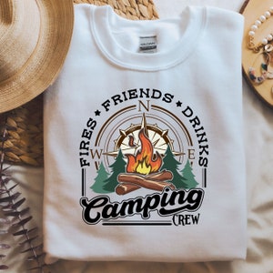 Könnte beinhalten: Weißer Pullover mit einem Kompass-Design mit einem Lagerfeuer, Bäumen und dem Text "Fires Friends Drinks Camping Crew".