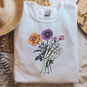 May include: White sweatshirt with a graphic of a skeleton hand holding a bouquet of flowers. The flowers are orange, purple, and pink. The text "For You" is written on the sweatshirt.