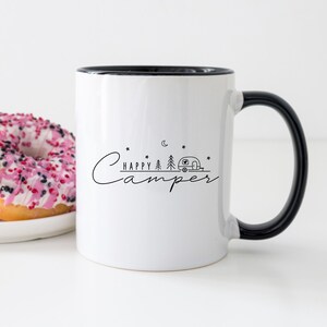 May include: White ceramic mug with a black rim and handle. The mug has a black graphic of a camper, trees, a moon, and stars with the text "Happy Camper" on it.