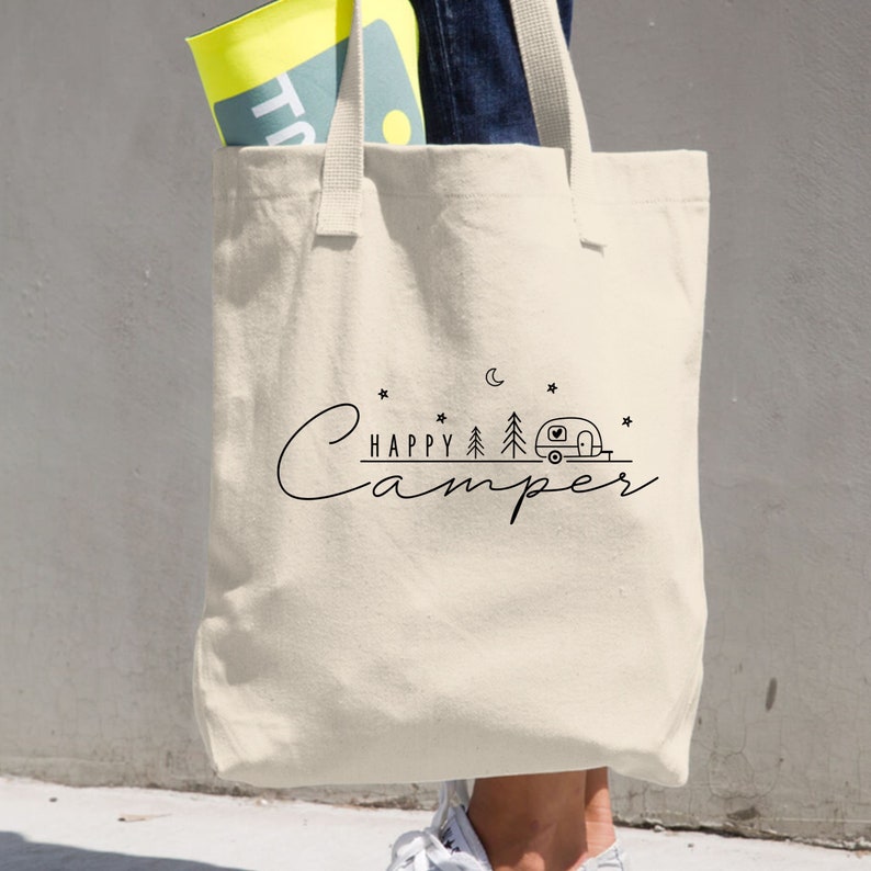 May include: A beige canvas tote bag with black text that reads "Happy Camper" and a small illustration of a camper van, trees, a crescent moon, and stars.