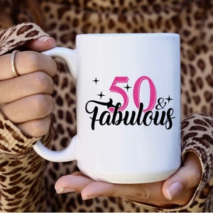 May include: White ceramic mug with a black and pink design that says "50 & Fabulous".
