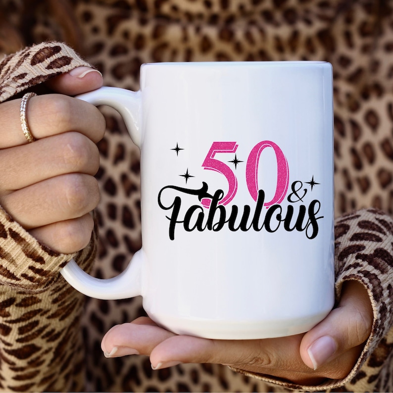May include: White ceramic mug with a black and pink design that says "50 & Fabulous".