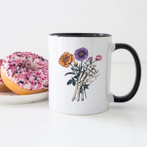 May include: White ceramic mug with a black rim and handle. The mug features a skeleton hand holding a bouquet of flowers, including an orange rose, a purple flower, and pink flowers. The text "For You" is written on the mug.