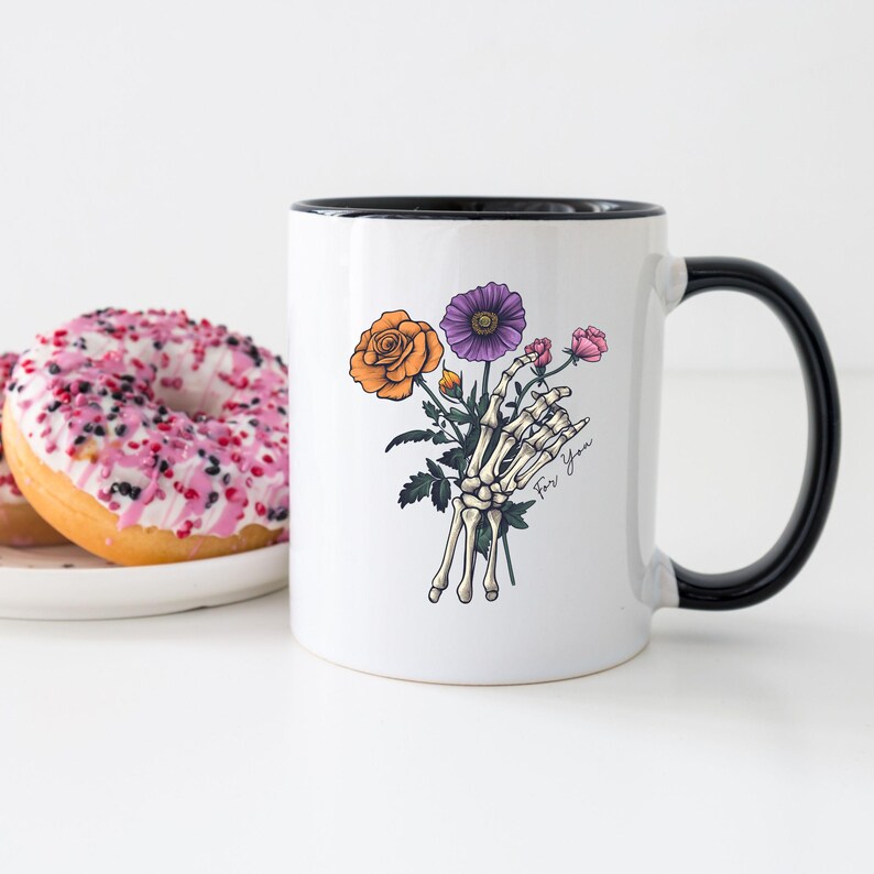 May include: White ceramic mug with a black rim and handle. The mug features a skeleton hand holding a bouquet of flowers, including an orange rose, a purple flower, and pink flowers. The text "For You" is written on the mug.