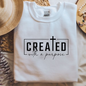 Könnte beinhalten: Weißer Pullover mit einem schwarzen Aufdruck, der "CREATED with a purpose" lautet und ein Kreuz über dem Wort "CREATED".