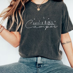 May include: A dark gray t-shirt with a white graphic design that says "Happy Camper" with a stylized camper, trees, and a crescent moon.