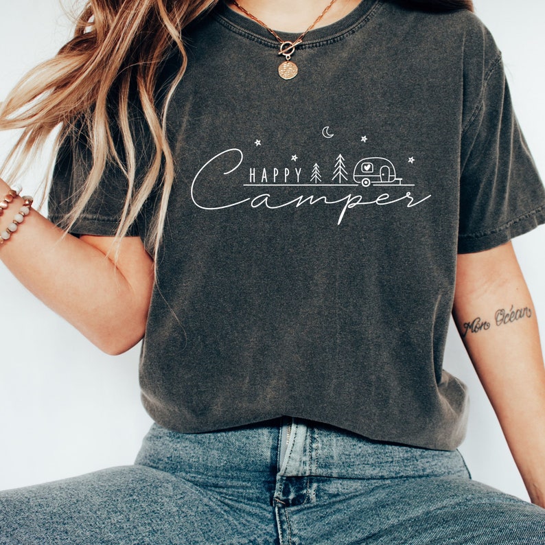 May include: A dark gray t-shirt with a white graphic design that says "Happy Camper" with a stylized camper, trees, and a crescent moon.
