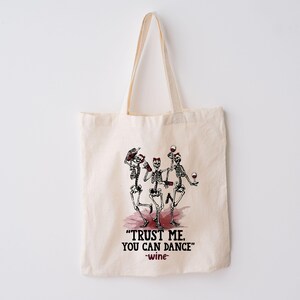 Peut inclure: Sac fourre-tout en coton beige avec un motif de trois squelettes dansant tenant des bouteilles et des verres de vin. Le texte "TRUST ME. YOU CAN DANCE" est en dessous, avec "-wine-".