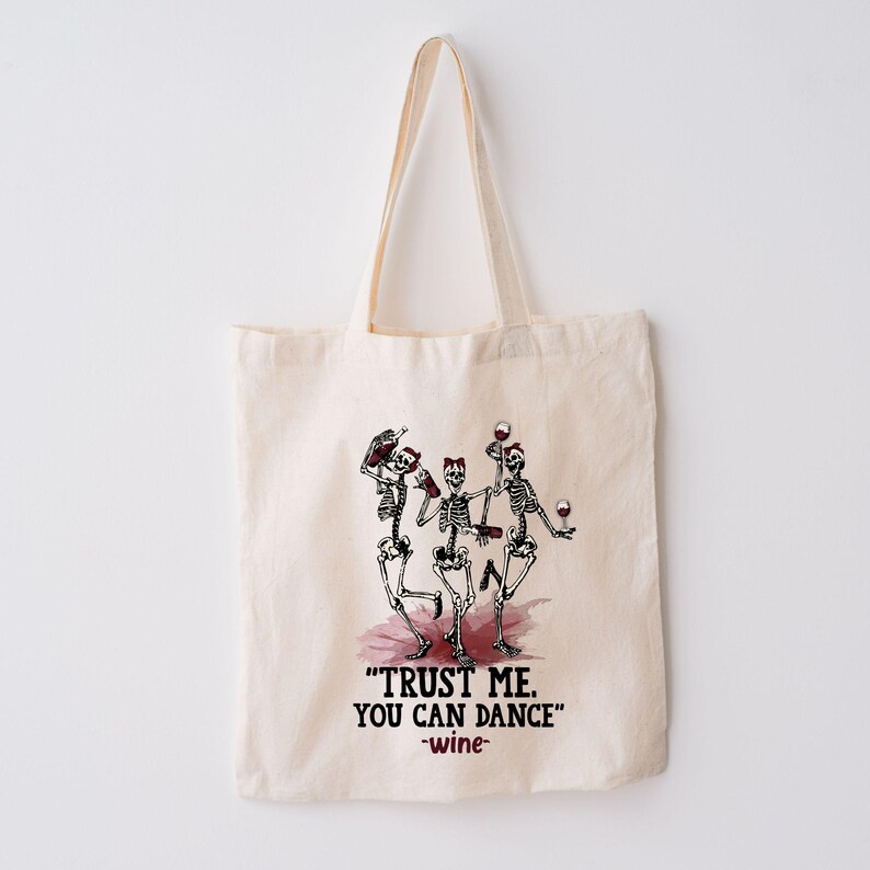 Peut inclure: Sac fourre-tout en coton beige avec un motif de trois squelettes dansant tenant des bouteilles et des verres de vin. Le texte "TRUST ME. YOU CAN DANCE" est en dessous, avec "-wine-".