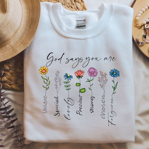 Puede incluir: Sudadera blanca con el texto "Dios dice que eres" y una fila de flores coloridas con palabras debajo de cada flor: "único", "especial", "adorable", "precioso", "fuerte", "elegido", "perdonado".